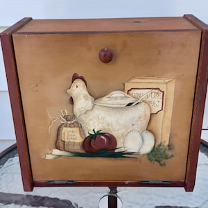 May include: Wooden bread box with a painted country scene. The artwork features a white hen, a red tomato, eggs, and a bag of herbs. The box has a brown knob and red trim.