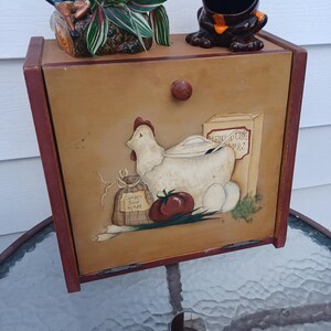 May include: A wooden storage box with a painted chicken and egg design, a frog-shaped container, and a small gnome figurine. The box has a brown knob and red trim. A small plant sits on top of the box.
