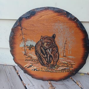 May include: A circular wooden plaque with a natural bark edge featuring a detailed illustration of a brown bear in a forest setting. The artwork depicts the bear centred among trees and sparse vegetation, rendered in shades of brown and tan.
