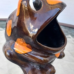 Peut inclure: Un récipient en céramique en forme de grenouille avec une large bouche ouverte. La grenouille est principalement marron avec des accents orange sur la tête et le corps. Les yeux sont blancs avec des pupilles noires. Le récipient est posé sur une table.