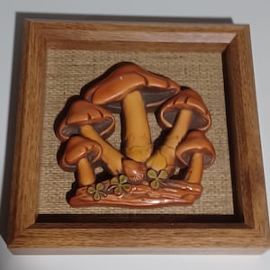 Peut inclure: Sculpture en relief encadrée représentant un groupe de champignons bruns et beiges. Les champignons sont disposés sur un fond texturé brun clair, dans un cadre en bois. L'œuvre comprend de petits trèfles verts.