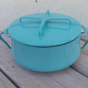 May include: A bright turquoise enameled metal casserole dish with a matching lid. The lid features a distinctive cross-shaped handle, and the pot has two curved side handles. The finish is smooth and glossy, covering the entire exterior surface.