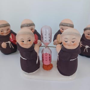 May include: A ceramic hourglass with pink sand, held by two monk figurines in brown robes. The hourglass is marked with numbers. Several other monk figurines surround the hourglass, all with smiling faces. The base is white.