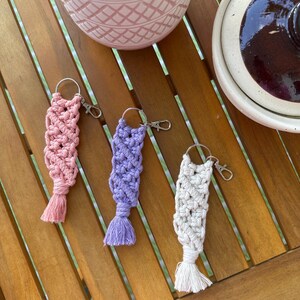 May include: Three macrame keychains in pink, purple, and white, each with a tassel and a silver-colored clasp. The keychains are displayed on a wooden surface, with a decorative pot and a ceramic jar in the background.