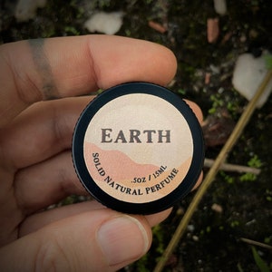 Close-up of Earth solid natural perfume tin in hand. A bold, earthy scent featuring vetiver, sandalwood, clary sage, & juniper. This herbaceous & smoky fragrance is perfect for those seeking an earthy aroma without patchouli.