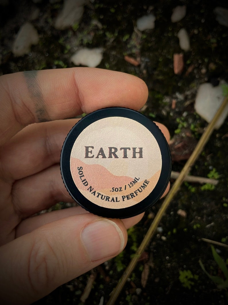 Close-up of Earth solid natural perfume tin in hand. A bold, earthy scent featuring vetiver, sandalwood, clary sage, & juniper. This herbaceous & smoky fragrance is perfect for those seeking an earthy aroma without patchouli.
