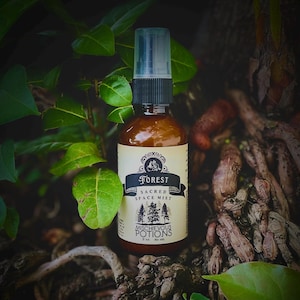 May include: A brown glass spray bottle labeled "Forest Sacred Space Mist" with a black and white label. The bottle is surrounded by green leaves and brown roots. The label includes the text "Mischievous Potions".