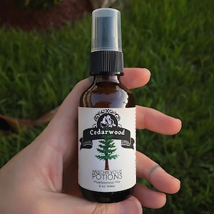 Puede incluir: Una botella de vidrio marrón con un pulverizador negro y una etiqueta blanca. La etiqueta dice "Cedarwood" y "Mischievous Potions Multifunctional Mist" con un dibujo de un cedro. La botella contiene 60 ml de líquido.
