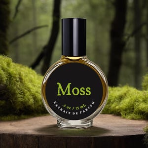Op de afbeelding: Een ronde glazen parfumfles met een zwarte dop en een zwart etiket met de groene tekst Moss en de witte tekst .5 oz / 15 ml Extrait de Parfum. De fles bevat een goudkleurige vloeistof en staat rechtop op een houten ondergrond.