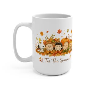 Könnte beinhalten: Ein weißer Keramikbecher mit einer farbenfrohen Illustration der Peanuts-Figuren, die in einem Haufen Herbstlaub liegen, zusammen mit einem Kürbis und einer Kiste Äpfel. Der Text Tis The Season ist in brauner Schrift unter den Figuren gedruckt.