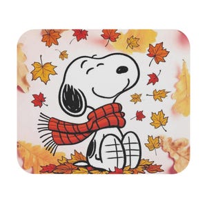 May include: A rectangular mouse pad featuring a cartoon illustration of a white beagle wearing a red fringed scarf. The character is surrounded by scattered autumn leaves in shades of orange, yellow, and red against a light background.