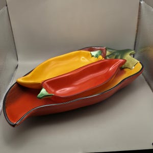 May include: A set of three ceramic serving dishes shaped like chili peppers. The largest is red, the medium is yellow, and the smallest is red. Each dish has a green stem and a black trim. A decorative and functional kitchen item.
