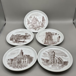 May include: Five white ceramic plates with brown architectural illustrations. Each plate features a different building or street scene, with text identifying the location. The plates are arranged on a gray surface.