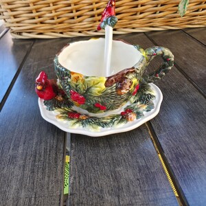 May include: A ceramic teacup and saucer set with a cardinal bird theme. The cup and saucer are decorated with green leaves, red berries, and a red cardinal. A white spoon with a cardinal on top rests in the cup. The set is on a wooden table.