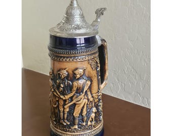 May include: A vintage beer stein with a detailed relief of figures in traditional attire, a dog, and grape vines. The ceramic stein has a brown and blue color scheme, a silver metal lid, and a handle.