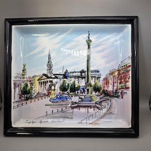 May include: A rectangular ceramic tray with a black frame, featuring a hand-painted scene of Trafalgar Square, London. The artwork includes buildings, a monument, and a blue sky with clouds. The tray is a decorative item.
