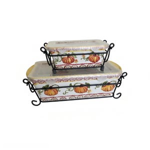 May include: Two rectangular ceramic baking dishes with clear lids, each nested in a black metal wire stand. The white dishes feature a decorative pattern of orange pumpkins, green vines, and yellow accents along the rims and base edges.