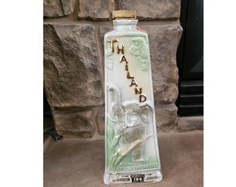 May include: A rectangular, off-white bottle with a gold cap and the word "THAILAND" in gold lettering. The bottle features a raised relief of an elephant and foliage. The words "AVIATION WONDERS" are at the bottom.