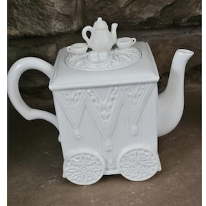 May include: A white ceramic teapot with a unique design, featuring a square body with decorative patterns and wheel-like accents. The lid is topped with a miniature teapot and two teacups. The teapot has a curved handle and a long spout.