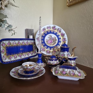 May include: A collection of fine porcelain tableware in a classic design. The set includes a serving tray, plates, teacups, a teapot, sugar bowl, creamer, and a butter dish. The pieces feature a royal blue and gold trim with a central image of a couple.