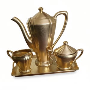 May include: A metallic gold tea set consisting of a tall teapot with a lid, a creamer, and a sugar bowl with a lid, all arranged on a matching rectangular serving tray. Each piece features a smooth, reflective finish and faceted sides.