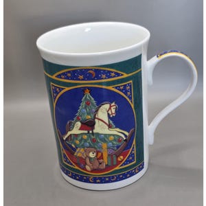 May include: White ceramic mug with a Christmas theme. The mug features a blue and green design with a Christmas tree, a rocking horse, and a teddy bear. The handle has a gold trim. The mug is approximately 4 inches tall.