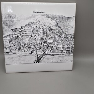 May include: A white ceramic tile featuring a black and white illustration of Heidelberg, Germany. The image depicts a detailed cityscape with the text "Heidelberga" and "Neckar Fluvius". The tile is rectangular.