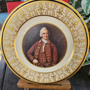 May include: A white porcelain plate with a gold rim and a blue border. The center of the plate features a portrait of a man in a red and gold jacket. The plate is on a wooden stand.