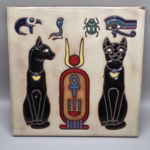 May include: Square ceramic tile featuring Egyptian motifs. The tile depicts two black cats, a central cartouche with an ankh symbol, and various hieroglyphic symbols in blue, green, and purple. The tile has a beige background and is likely a decorative item.