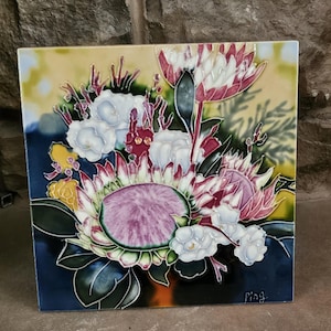 May include: A ceramic tile with a colorful floral design featuring pink, white, and green protea flowers. The tile has a blue background and a yellow border. The tile is signed "Ping" in the bottom right corner.