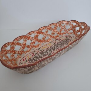 May include: An elongated ceramic basket with a scalloped rim and an intricate lattice-style openwork design. The exterior features a repeating orange floral pattern, while the interior base displays a central band of dark brown geometric motifs.