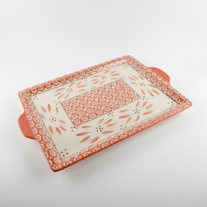 May include: A rectangular ceramic serving tray with integrated handles, featuring a cream-colored center and a terracotta-orange border. The design includes a central panel of repeating floral patterns surrounded by stylized leaf motifs.