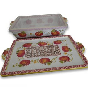 May include: A rectangular ceramic baking dish with a matching lid, both featuring a white base decorated with orange pumpkins, green vines, and a red and yellow floral border. The lid has a central rectangular pattern of small brown flowers.