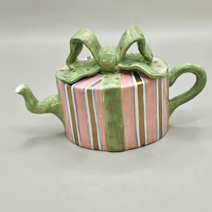 May include: A ceramic teapot shaped like a gift box, featuring pink and gold vertical stripes. The lid is topped with a green bow with gold polka dots. The spout and handle are also green.
