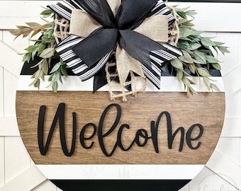 May include: A round wooden door sign featuring horizontal stripes in black, white, and natural wood tones. The word Welcome is written in black script lettering. A large decorative bow made of black, white striped, and burlap ribbon sits on top.