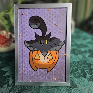 May include: A framed illustration of a black and orange pumpkin with a black cat-like face and a curled tail. The background is purple with white polka dots and orange candy corn.