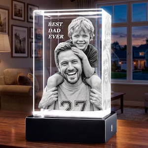 May include: A rectangular glass block featuring a laser-etched black and white photograph of a child sitting on a person's shoulders. The text "BEST DAD EVER" is etched in the upper left corner. The block sits on a solid black rectangular base.