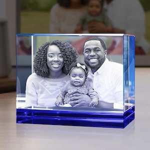 May include: A rectangular glass block featuring a laser-etched black and white photograph of a family consisting of a person with curly hair, a person with a beard, and a baby wearing a small bow. The base of the glass block is deep blue.