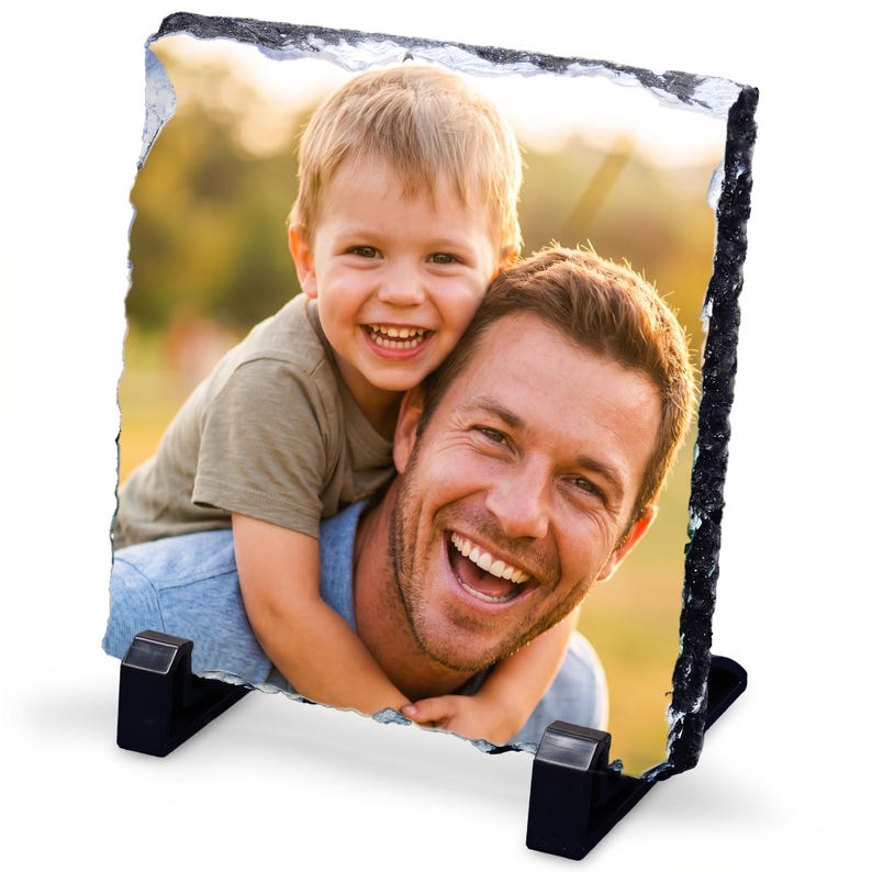 May include: A square-shaped photo slate with rough, chiseled edges featuring a color photograph of a smiling child hugging a smiling adult. The slate is supported by two black plastic stands positioned at the bottom corners of the stone plaque.