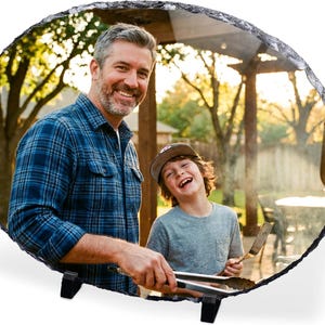 May include: An oval-shaped slate photo plaque featuring a printed image of an adult and a child grilling outdoors. The plaque has a rough, natural stone edge and is supported by two small black plastic stands on a plain white background.
