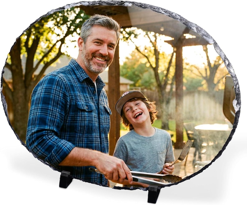 May include: An oval-shaped slate photo plaque featuring a printed image of an adult and a child grilling outdoors. The plaque has a rough, natural stone edge and is supported by two small black plastic stands on a plain white background.