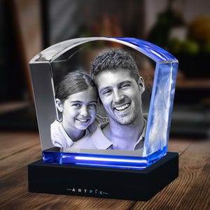 May include: A clear, arch-topped glass block featuring a laser-etched black and white photograph of a smiling child and adult. The block sits on a black rectangular base with the text ARTPIX printed in white, illuminated by a blue light.