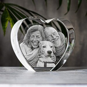 Puede incluir: Un bloque de cristal transparente en forma de coraz&oacute;n con una fotograf&iacute;a en blanco y negro grabada con l&aacute;ser de dos personas sonrientes y un perro. El cristal es grueso y transparente con bordes pulidos, mostrando el retrato detallado.