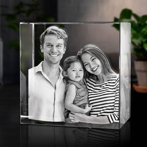 May include: A clear, rectangular glass block featuring a laser-etched, black and white photographic portrait of a family. The image displays a smiling adult wearing a collared shirt, a young child, and another smiling adult in a striped top.