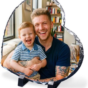 May include: A heart-shaped slate photo plaque featuring a printed image of a smiling adult and a young child. The plaque has a rough, natural stone edge and is supported by two black plastic stands positioned at the base of the heart shape.