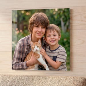 May include: A square glass wall art print featuring a photograph of two smiling children holding a ginger and white cat. The older child wears a brown plaid shirt, while the younger child wears a horizontal striped t-shirt with a chest pocket.