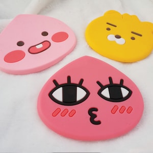 May include: Three silicone coasters featuring cartoon characters. The first coaster is pink and shaped like a peach with a smiling face. The second coaster is yellow and shaped like a bear with a frowning face. The third coaster is pink and shaped like a peach with large eyes and a blushing face.