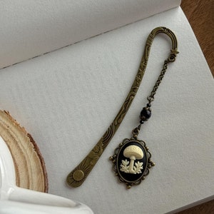 Puede incluir: Un marcapáginas de bronce con un diseño detallado que presenta estrellas y una luna creciente. Una cadena negra con una cuenta negra cuelga del marcapáginas y se conecta a un camafeo que presenta un hongo blanco sobre un fondo negro.