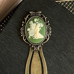 May include: Antique bronze bookmark with an oval cameo featuring a fairy in cream against a green background. The bookmark is placed on a black book, with a partially visible open book and decorative artwork in the background.