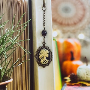 May include: A vintage-style bookmark with a cameo pendant featuring a skeleton design. The pendant is attached to a chain and a clasp. The bookmark is hanging from a book. The background includes a plant, pumpkins, and a book.